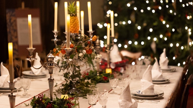 Festive dining table with cone-shaped napkins, crystal glasses, and a candelabrum topped with a pineapple, set against a lit Christmas tree.